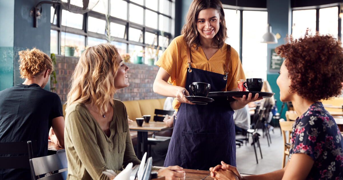 Zwei Freundinnen sitzen am Tisch im Coffee Shop und werden von der Kellnerin bedient